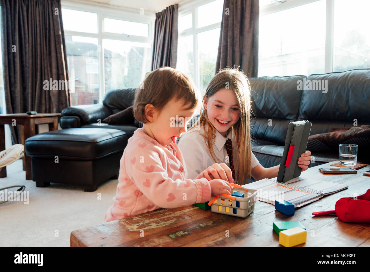 Little girl is distracting her older sister while she is trying to study at home. They are playing with toy blocks together. Stock Photo