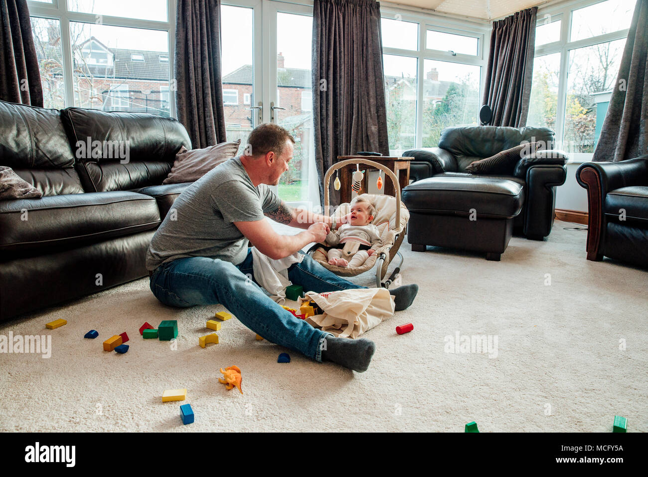Stay at home father is relaxing in the living room with his baby daughter while she is in her baby bouncer. Stock Photo