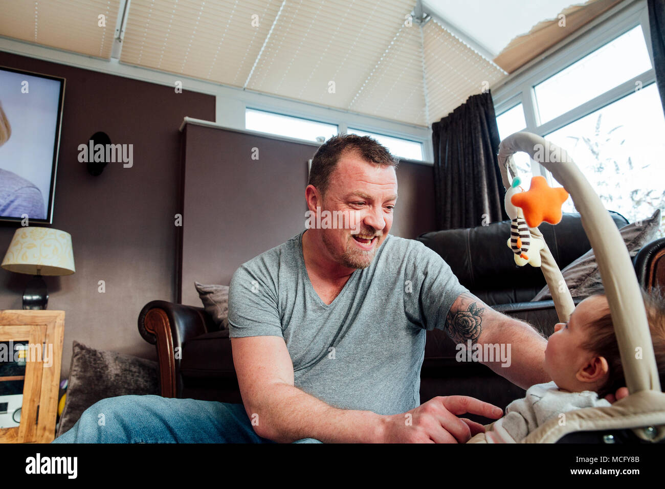 Stay at home father is relaxing in the living room with his baby daughter while she is in her baby bouncer. Stock Photo