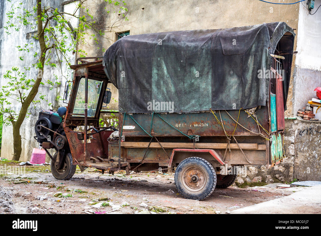 An old three wheeled truck with exposed engine and drive belt. The rear of the truck is covered with a tarpaulin. Daxu, China. Stock Photo