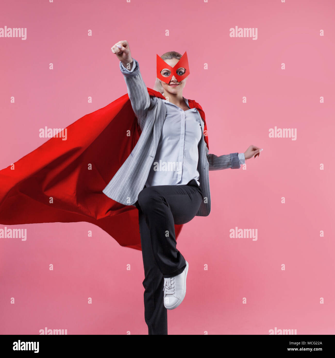 Young attractive woman superhero. Girl in a business suit and a mask with red cloak of hero. On a pink background. Jumps Stock Photo