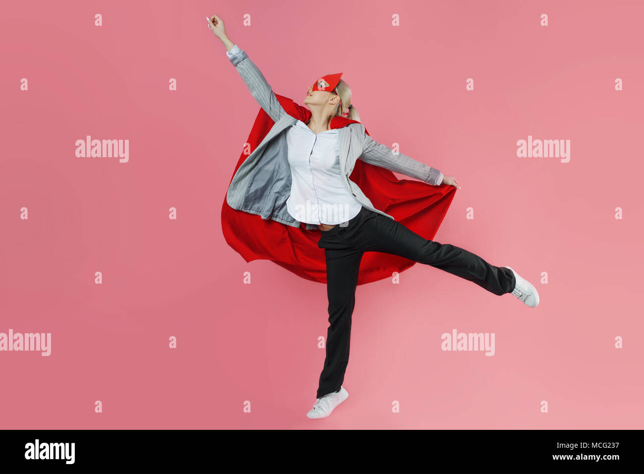 Young attractive woman superhero. Girl in a business suit and a mask with red cloak of hero. On a pink background. In flight, arm up Stock Photo