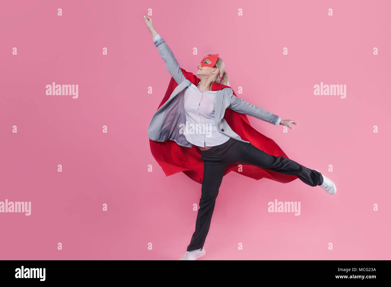 Young attractive woman superhero. Girl in a business suit and a mask with red cloak of hero. On a pink background. In flight, arm up Stock Photo