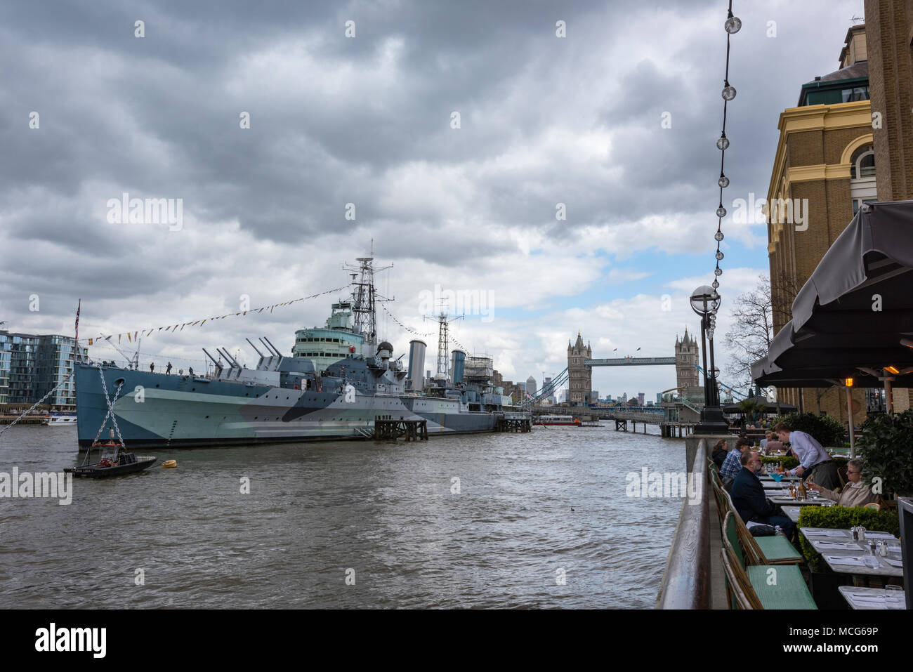 HMS Belfast alongside on the river thames in central london with people eating at a restaurant on the south bank riverside on a warm but overcast day. Stock Photo