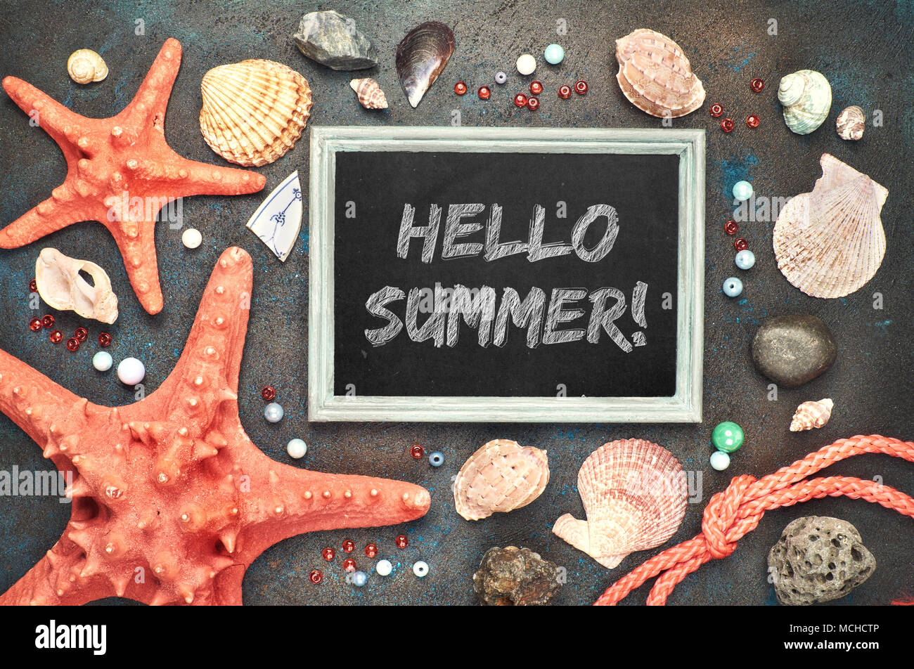Blackboard with 'Hello Summer!' chalk text, with sea shells, rope and star fish on dark background Stock Photo
