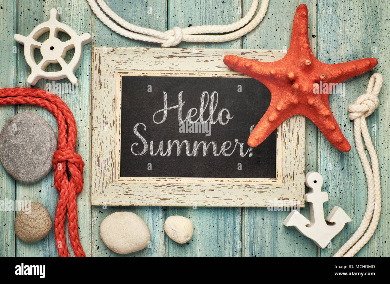 Blackboard with 'Hello Summer!' chalk text, with sea shells, rope and star fish on light green wooden background Stock Photo