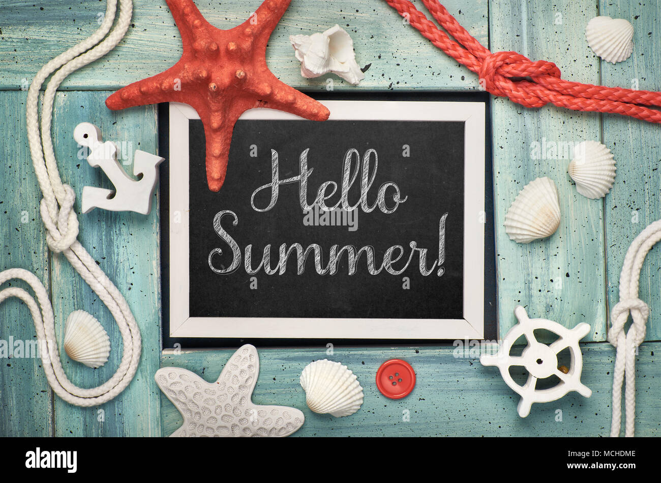 Blackboard with 'Hello Summer!' chalk text, with sea shells, rope and star fish on light green wooden background Stock Photo