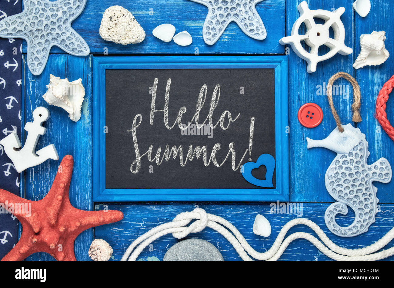 Blackboard with 'Hello Summer!' chalk text, with sea shells, stones, rope and star fish on blue wooden background Stock Photo