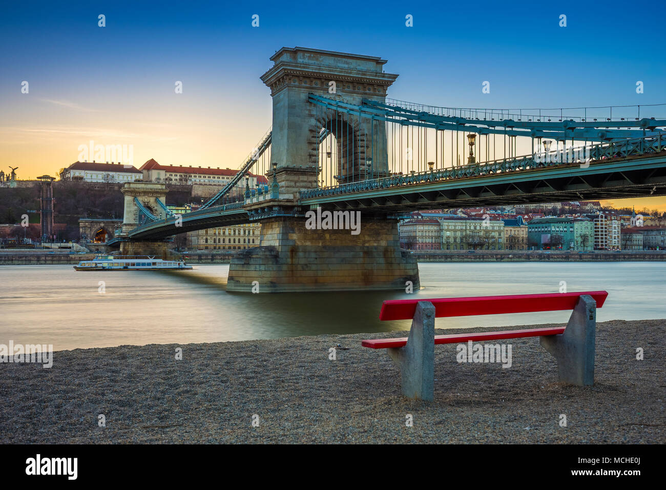 Budapest, Hungary - Famous Szechenyi Chain Bridge with traditional red bench and sightseeing boat on River Danube at sunset. Clear blue sky Stock Photo