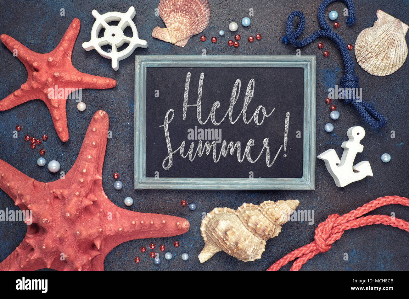 Blackboard with 'Hello Summer!' chalk text, with sea shells, rope and star fish on dark blue background Stock Photo
