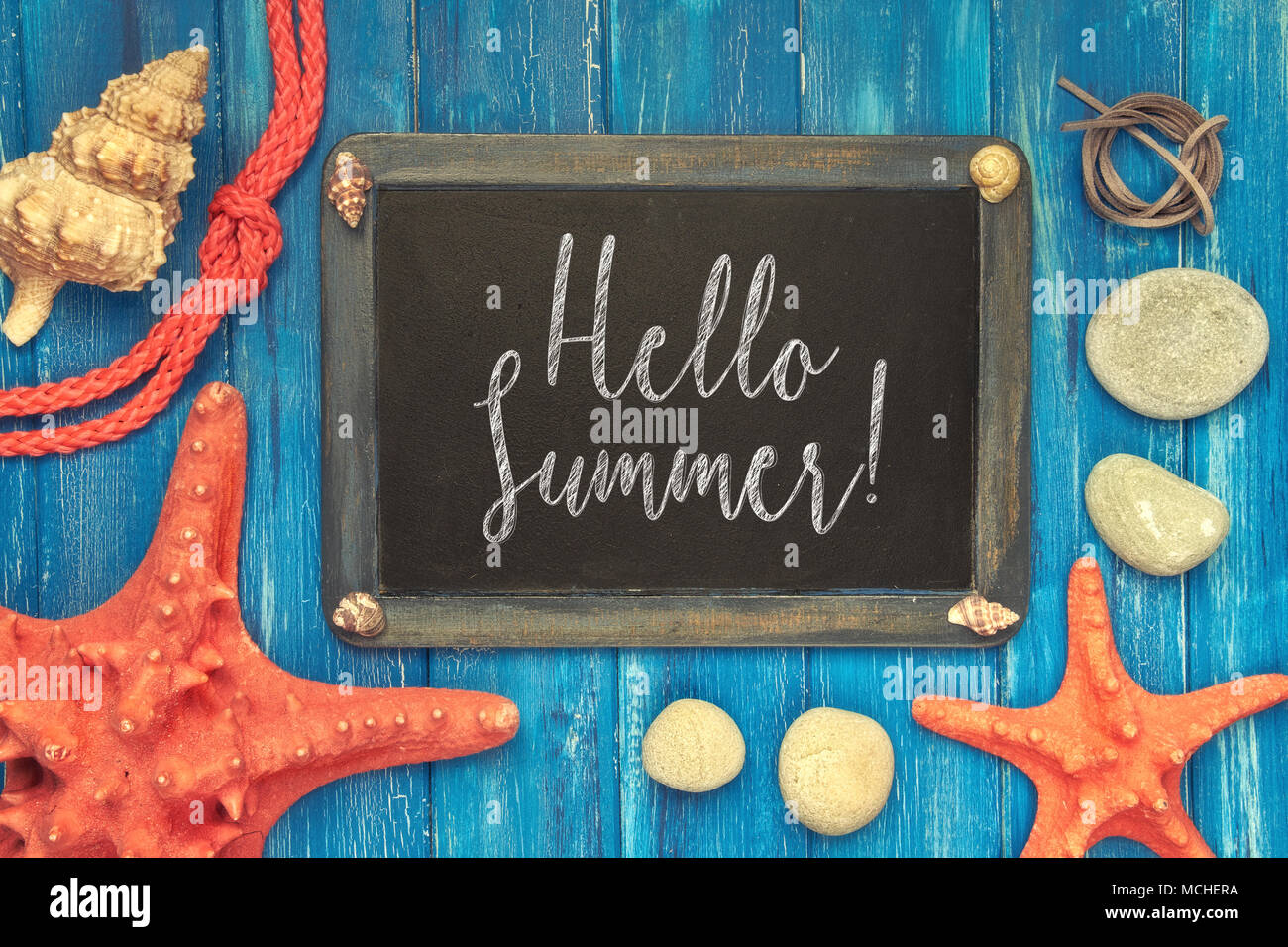Blackboard with 'Hello Summer' chalk text, with sea shells, rope and star fish on blue  wooden background Stock Photo