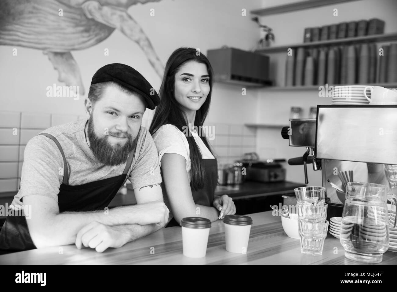 Coffee Business Concept - Positive young bearded man and beautiful attractive lady barista couple in apron looking at camera while standing at bar Counter Stock Photo