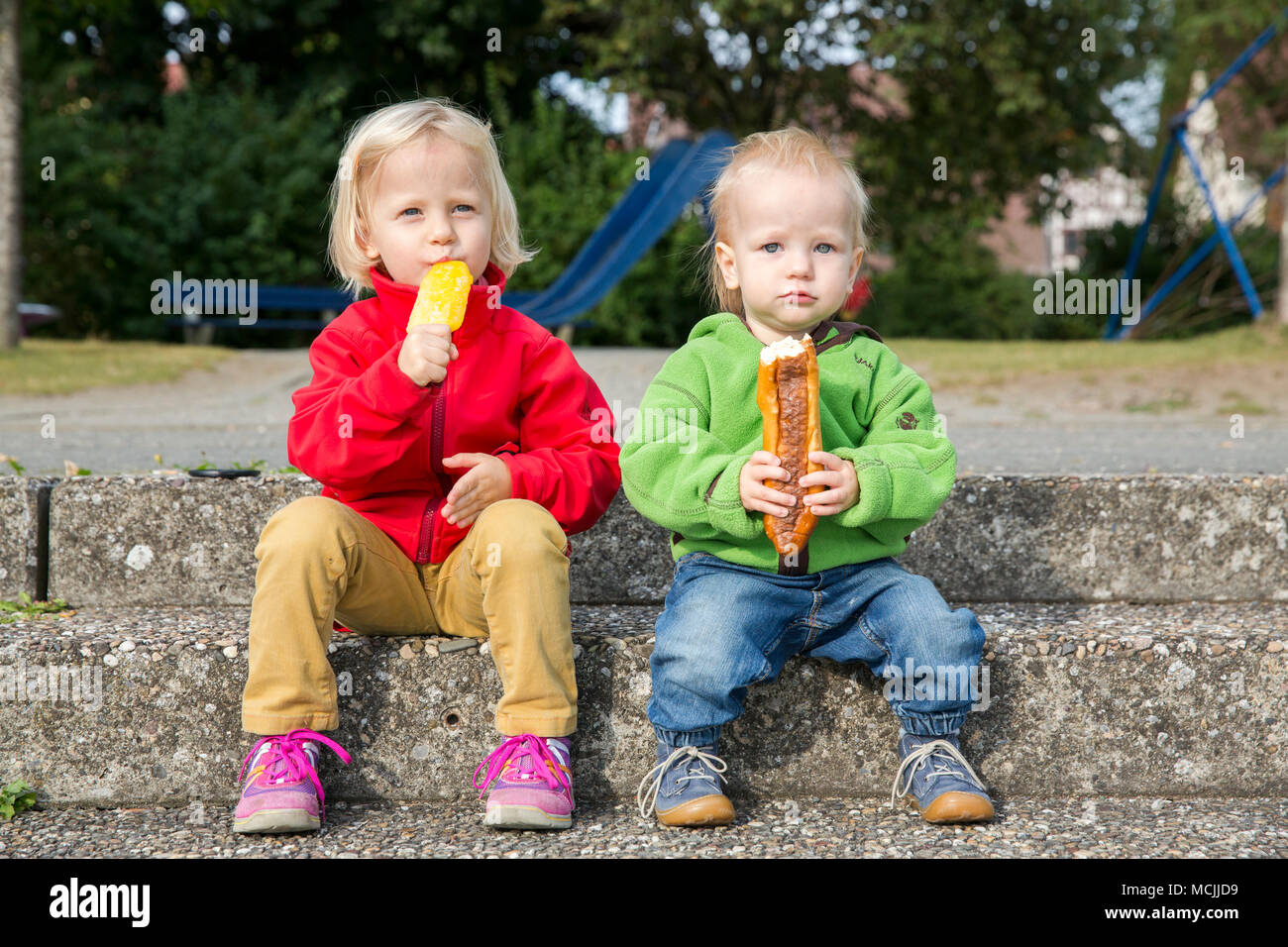 Infants, siblings, sitting on stairs eating ice cream and pretzel sticks Stock Photo