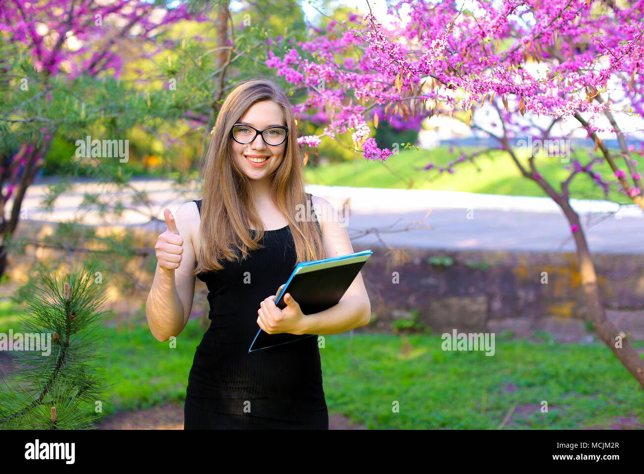 Cute female student showing thumbs up in garden with documents in black ...