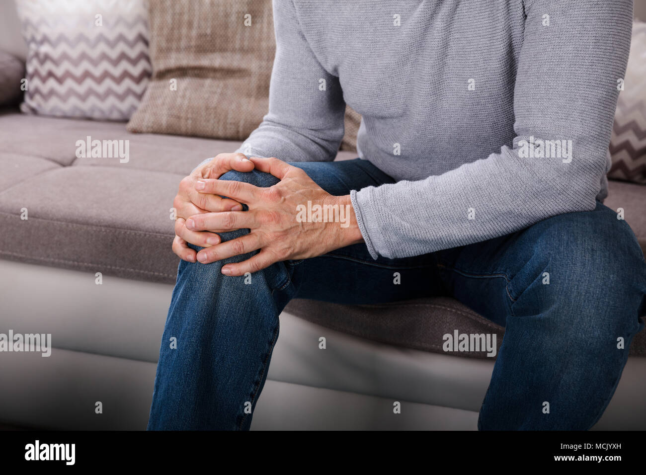 Man Suffering From Knee Pain Sitting On Sofa Stock Photo