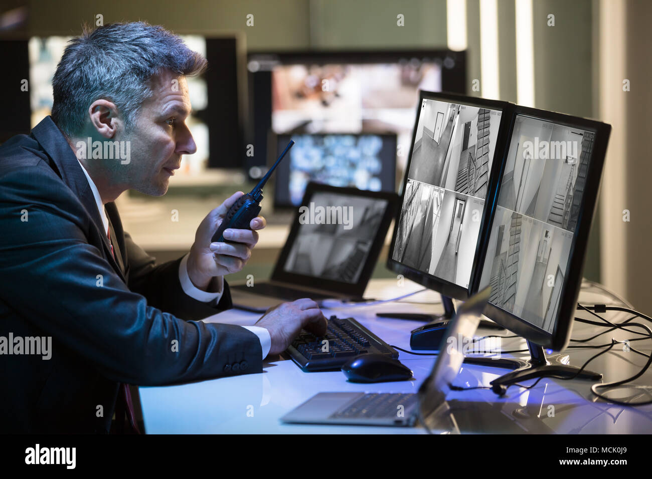 Businessman Talking On Walkie Talkie While Looking At CCTV Camera Footage On Multiple Computer Screen Stock Photo