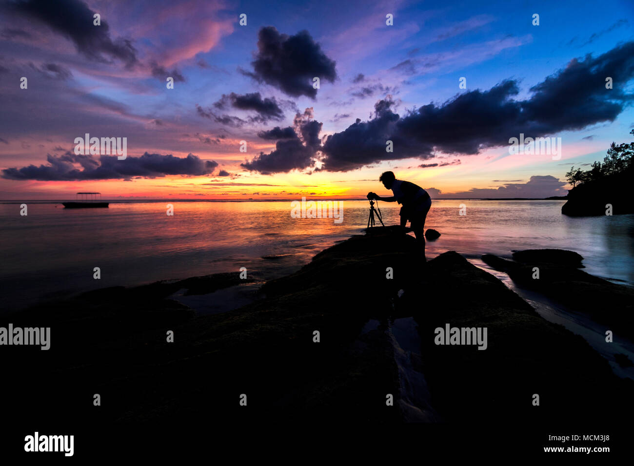 Silhouette of photographer taking photos of a dramatic sunset with tripod on the rock. Stock Photo