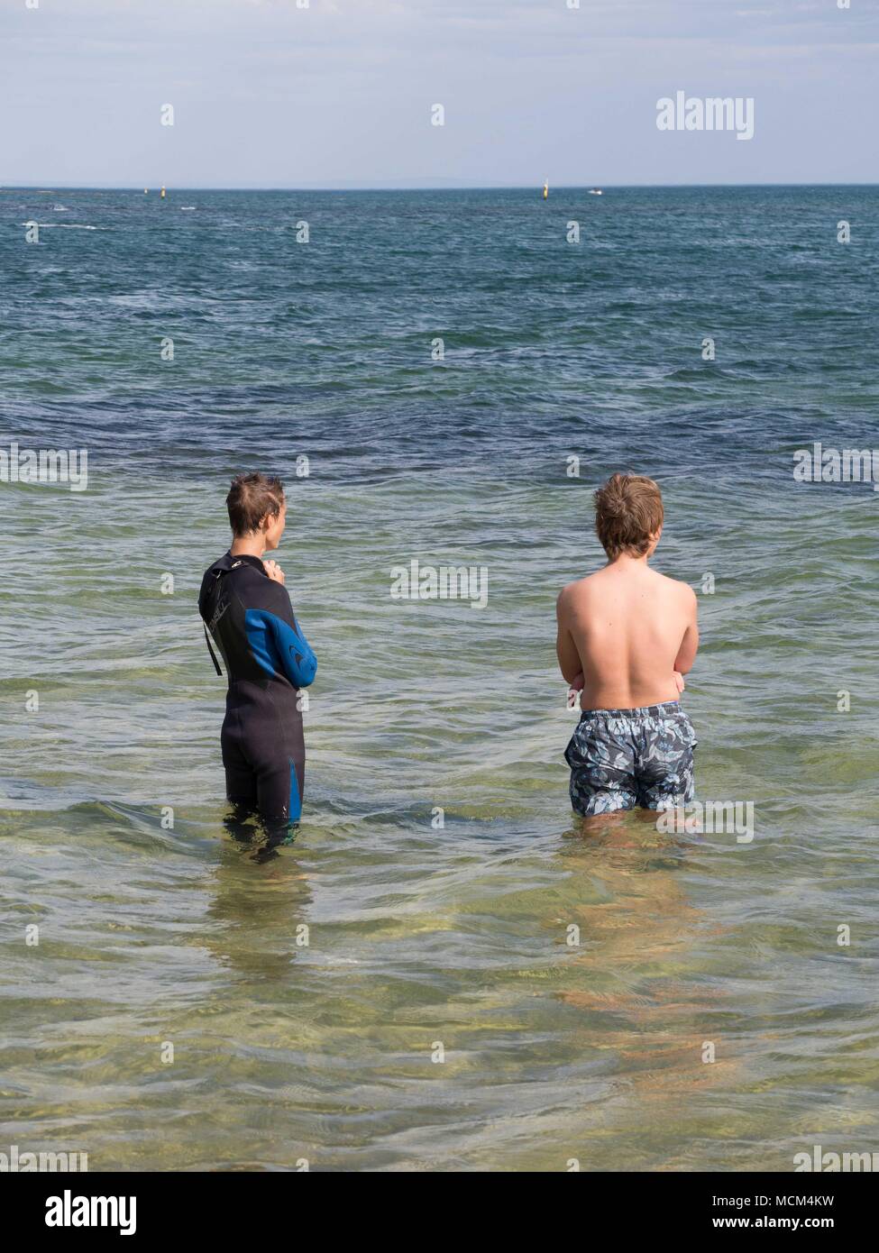 Two boys standing waist deep in water one in a wetsuit other in bathing