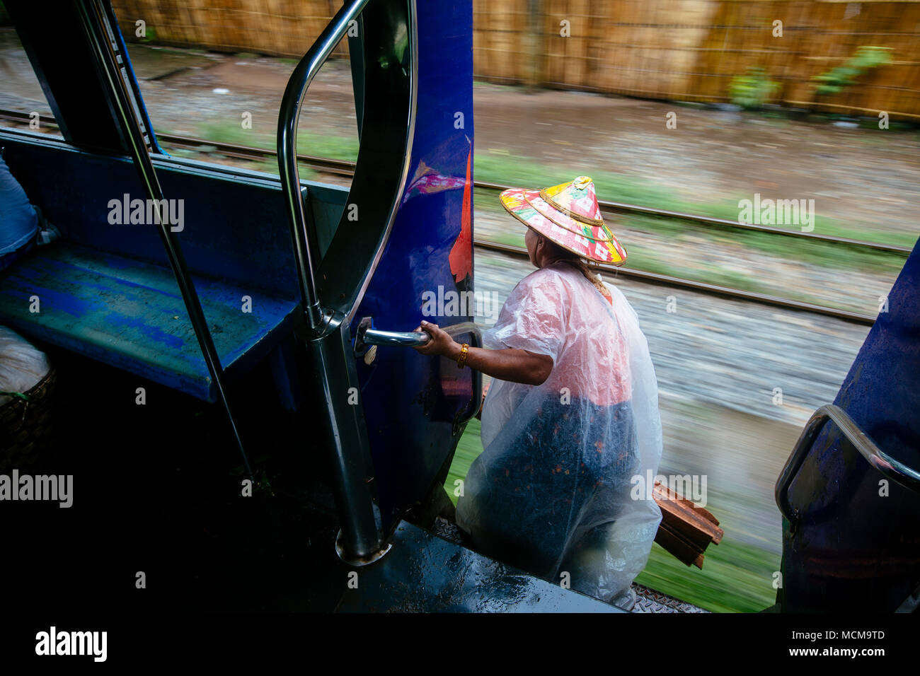 Woman wearing traditional hat and plastic raincoat leaning out of moving train, Yangon, Myanmar Stock Photo
