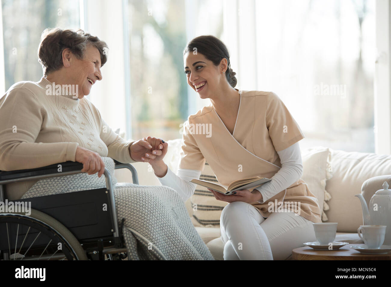 Senior in a wheelchair and beautiful nurse smiling and holding hands while reading a book Stock Photo