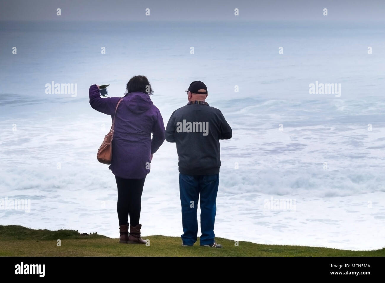 UK weather - A couple standing on the coast in misty weather overlooking the sea and taking photographs of the sea with a smartphone. Stock Photo