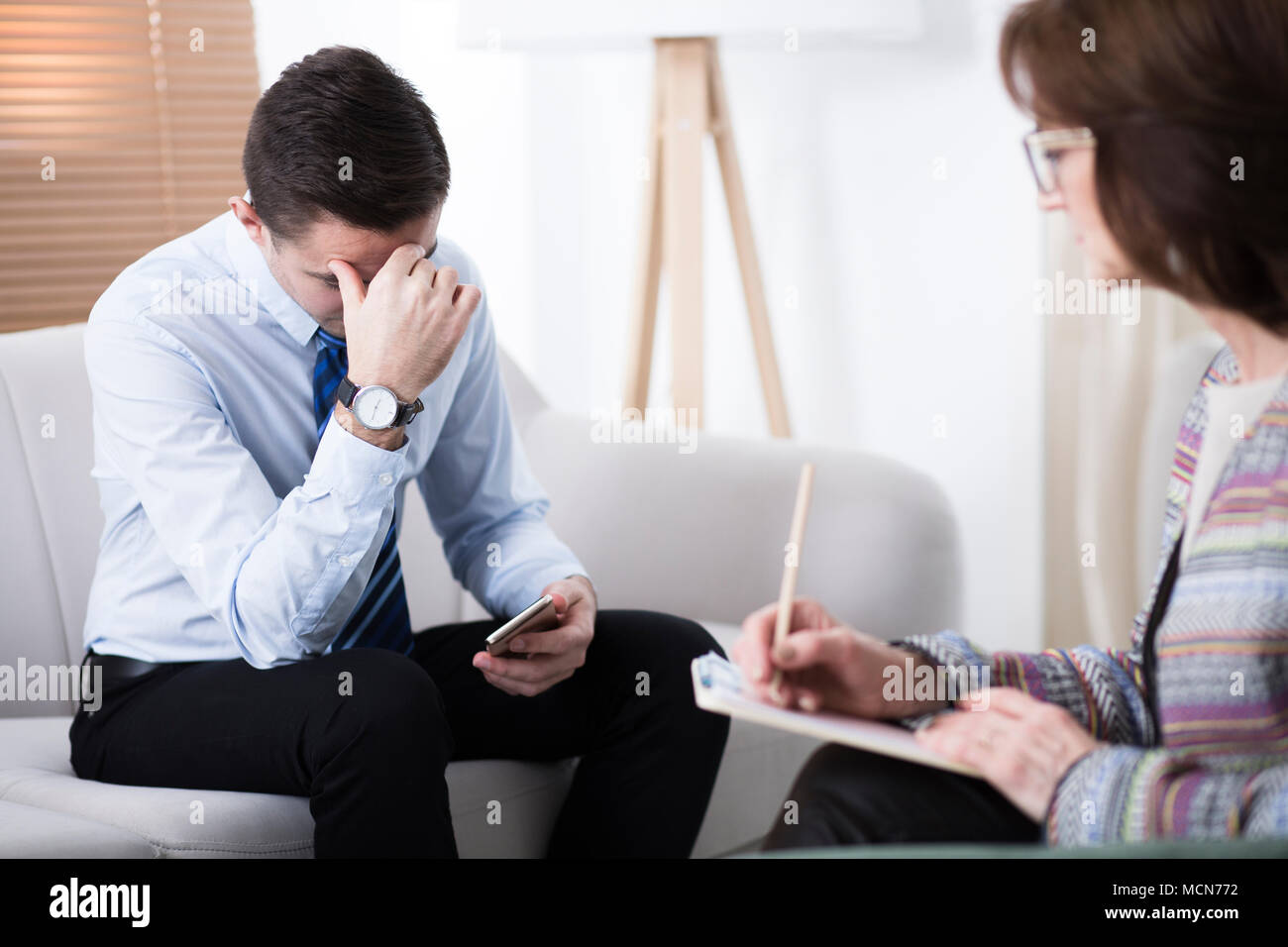 Depressed man with problems looking at his phone while sitting in a psychotherapist's office Stock Photo