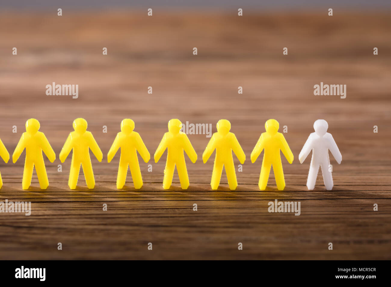 White Figure Standing Among Yellow Human Figures In A Row On Wooden Background Stock Photo
