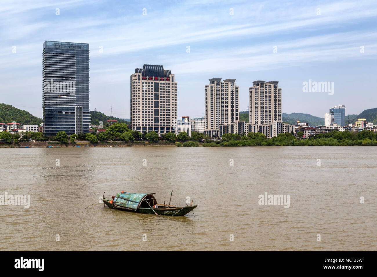 A sampan floats on the Fuchun River juxtaposed by a number of modern high rise buildings along the riverbank. Tonglu, Zhejiang Province, China. Stock Photo