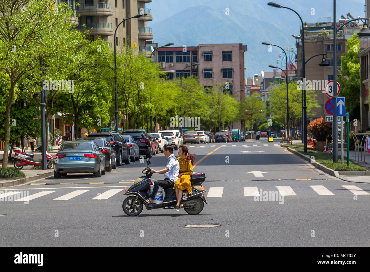 A young Chinese couple ride on a motor scooter. The girl wears a yellow dress and rides pillion sitting side saddle. Tonglu, Zhejiang Province, China. Stock Photo