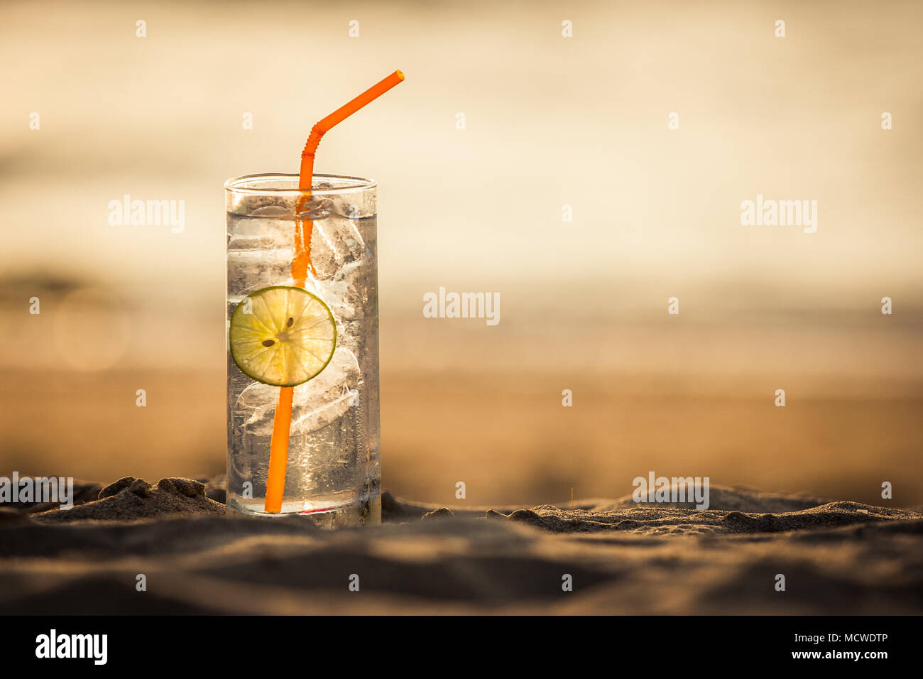 Picture of a glass of Gin Tonic with straw and lime slice on the beach, at sunset. Long Beach, Ko Lanta, Thailand. Stock Photo