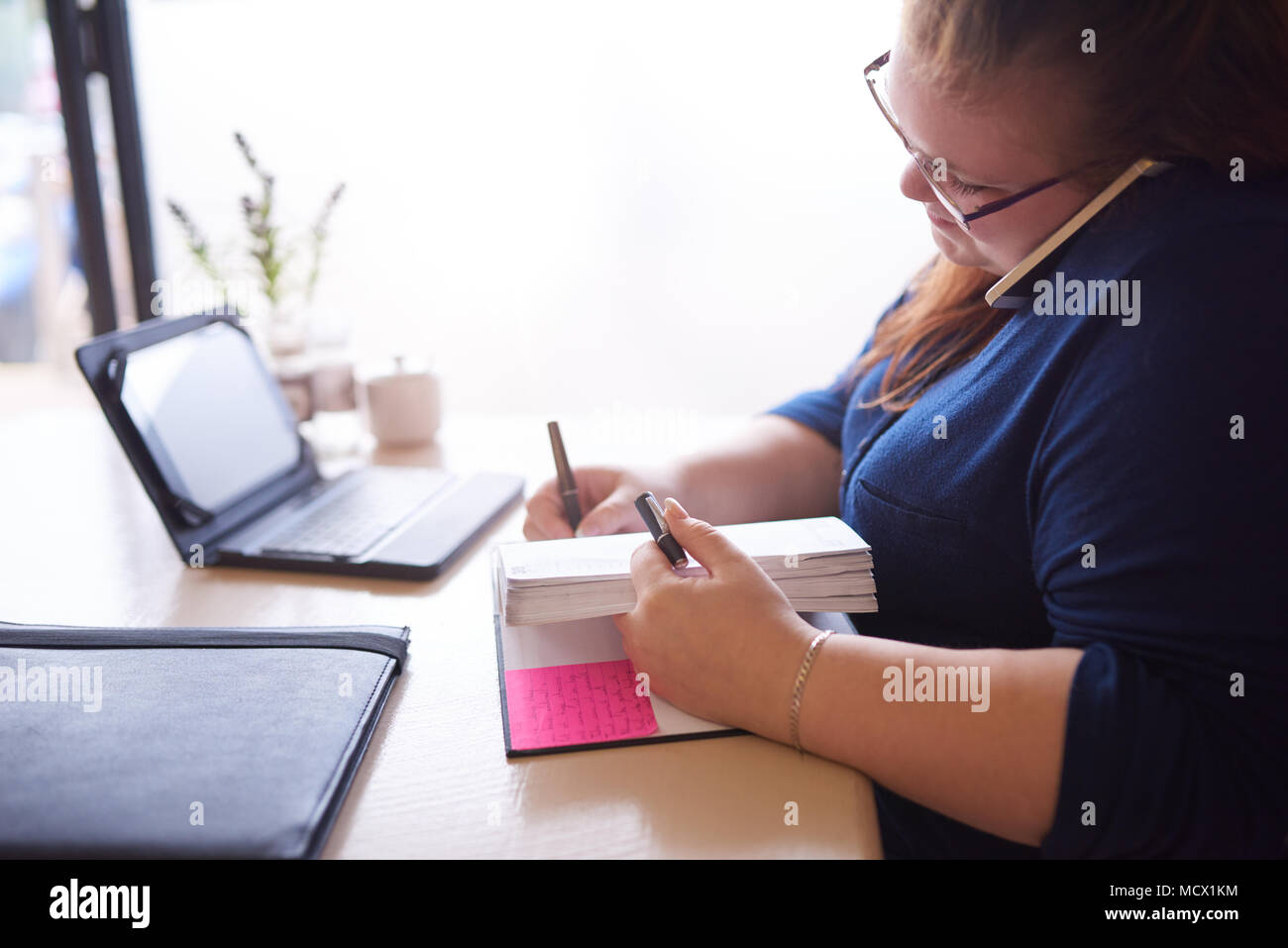 Moderately overweight caucasian woman seated at her desk busy multitasking while she writes in her notebook and talks on her phone, that is pinched between her head and shoulders. Stock Photo