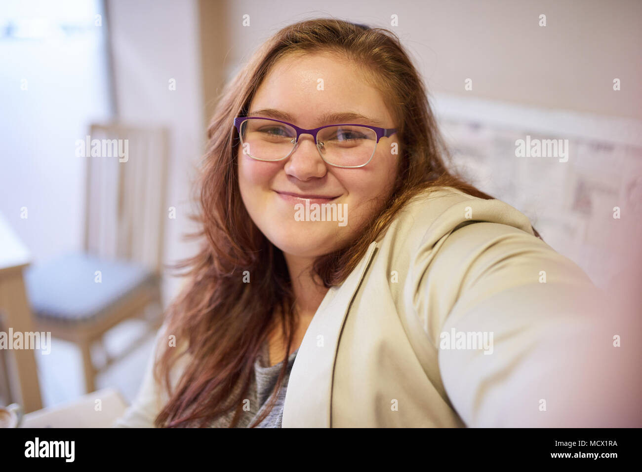 Slightly overweight beautiful young caucasian woman taking a picture of herself while wearing formal clothing and glasses in a cafe to start her day off with a smile and positive energy. Stock Photo