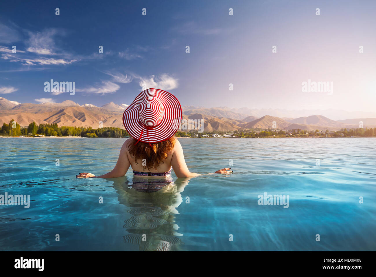 Beautiful woman in red striped hat swimming in crystal clean Issyk Kul lake with mountains background at sunrise in Kyrgyzstan Stock Photo