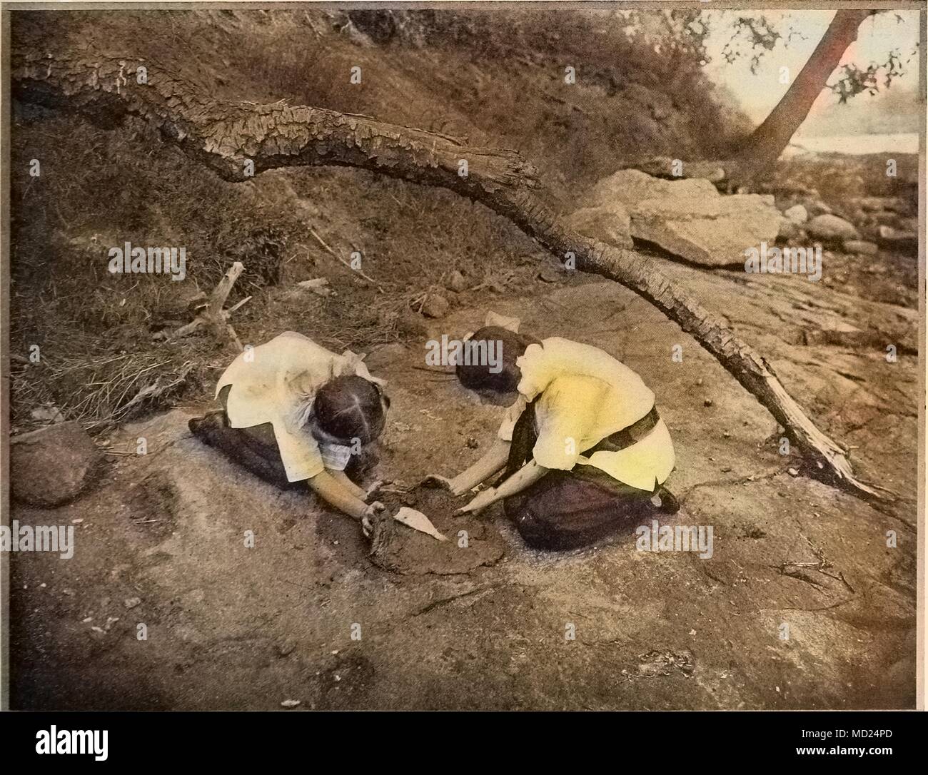 Black and white photograph of two women outdoors, preparing a fish for baking by rubbing soft clay on it, 1905. Courtesy Internet Archive. Note: Image has been digitally colorized using a modern process. Colors may not be period-accurate. () Stock Photo