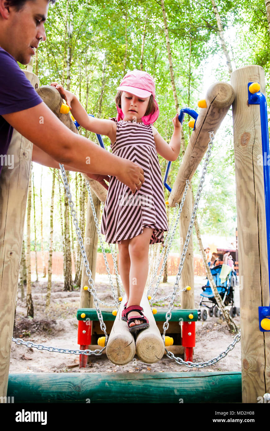 Father helping her daughter cross balance beam on playground, spending time together. Stock Photo