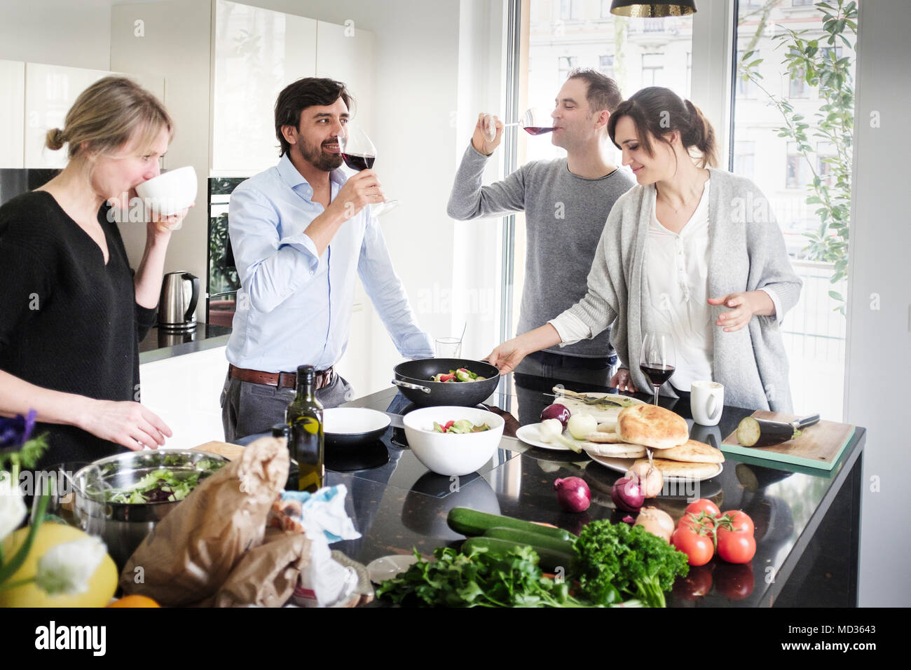 Group of friends  cooking together while casuallyhaving a drink and  snacking on a selection of vegetarian  food. Laughing and enjoying themselves. Stock Photo