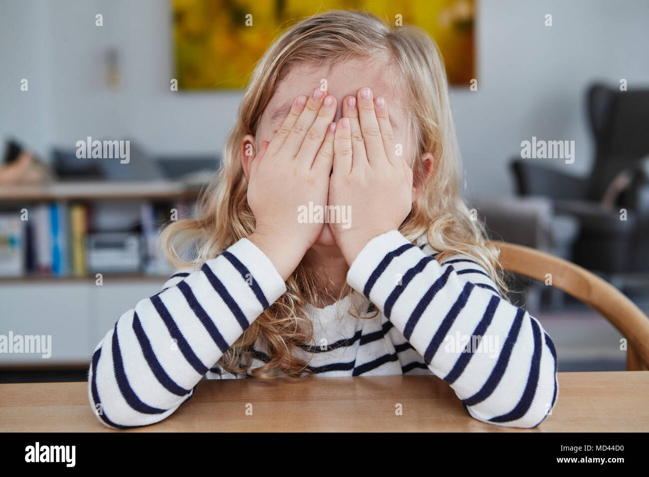 Portrait of young girl, sitting at table, covering face with hands Stock Photo