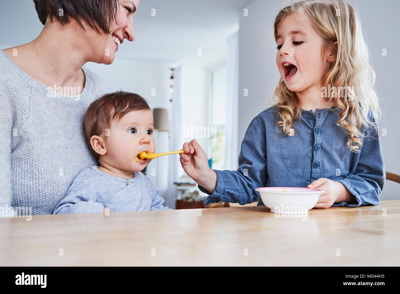 Family sitting at kitchen table, young girl spoon-feeding baby sister Stock Photo