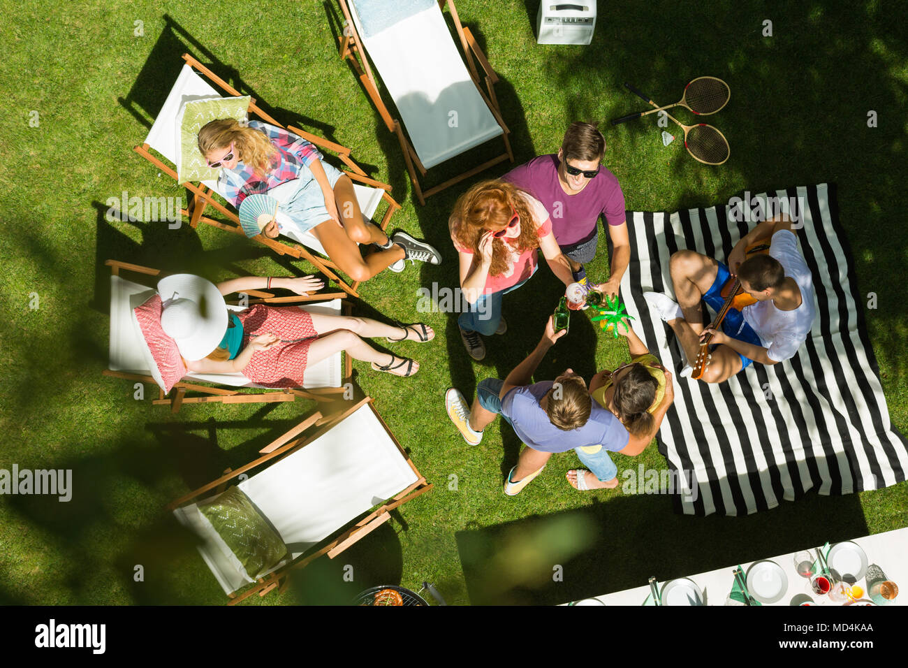 Top view of group of friends enjoying summertime garden party in the countryside, sitting on deck chairs, talking and sunbathing surrounded by nature Stock Photo