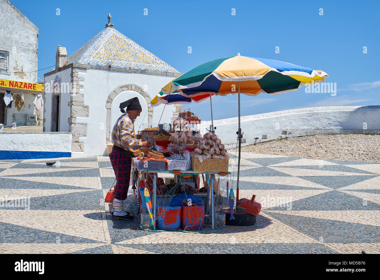 NAZARE, PORTUGAL - JUNE 26, 2016: The street vendor in the colorful traditional costume on the central square of Nazare with the Memory Chapel (Ermida Stock Photo