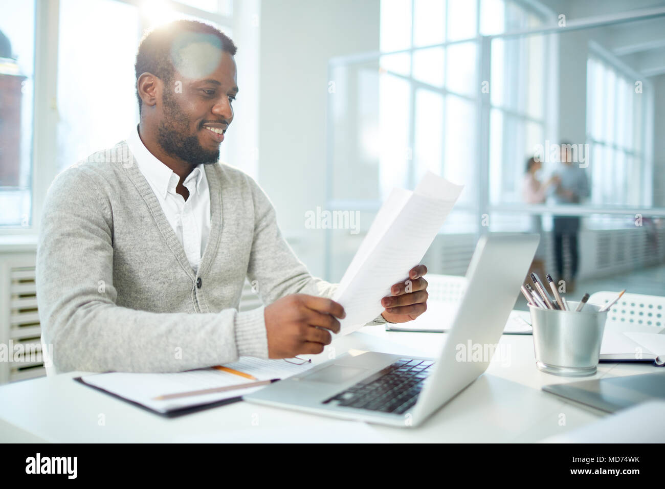 Waist-up portrait of cheerful African American businessman studying document while sitting at desk of modern open plan office, lens flare Stock Photo