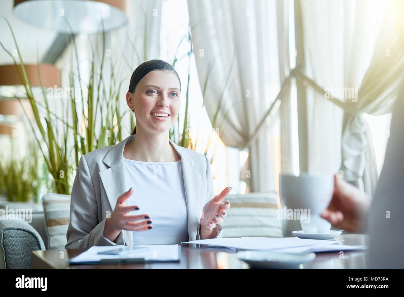 Young attractive Caucasian businesswoman discussing project with colleague and smiling happily during working lunch in cafe Stock Photo