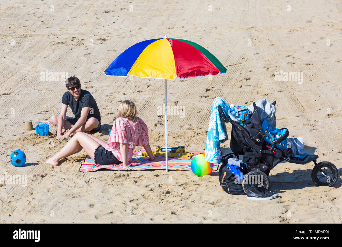 Bournemouth, Dorset, UK. 19th April 2018. UK weather: lovely warm sunny day at Bournemouth beaches with clear blue skies and unbroken sunshine, as visitors head to the seaside to enjoy the warmest day of the year so far. Couple relaxing on the beach under colourful parasol with pushchair buggy by the side. Credit: Carolyn Jenkins/Alamy Live News Stock Photo