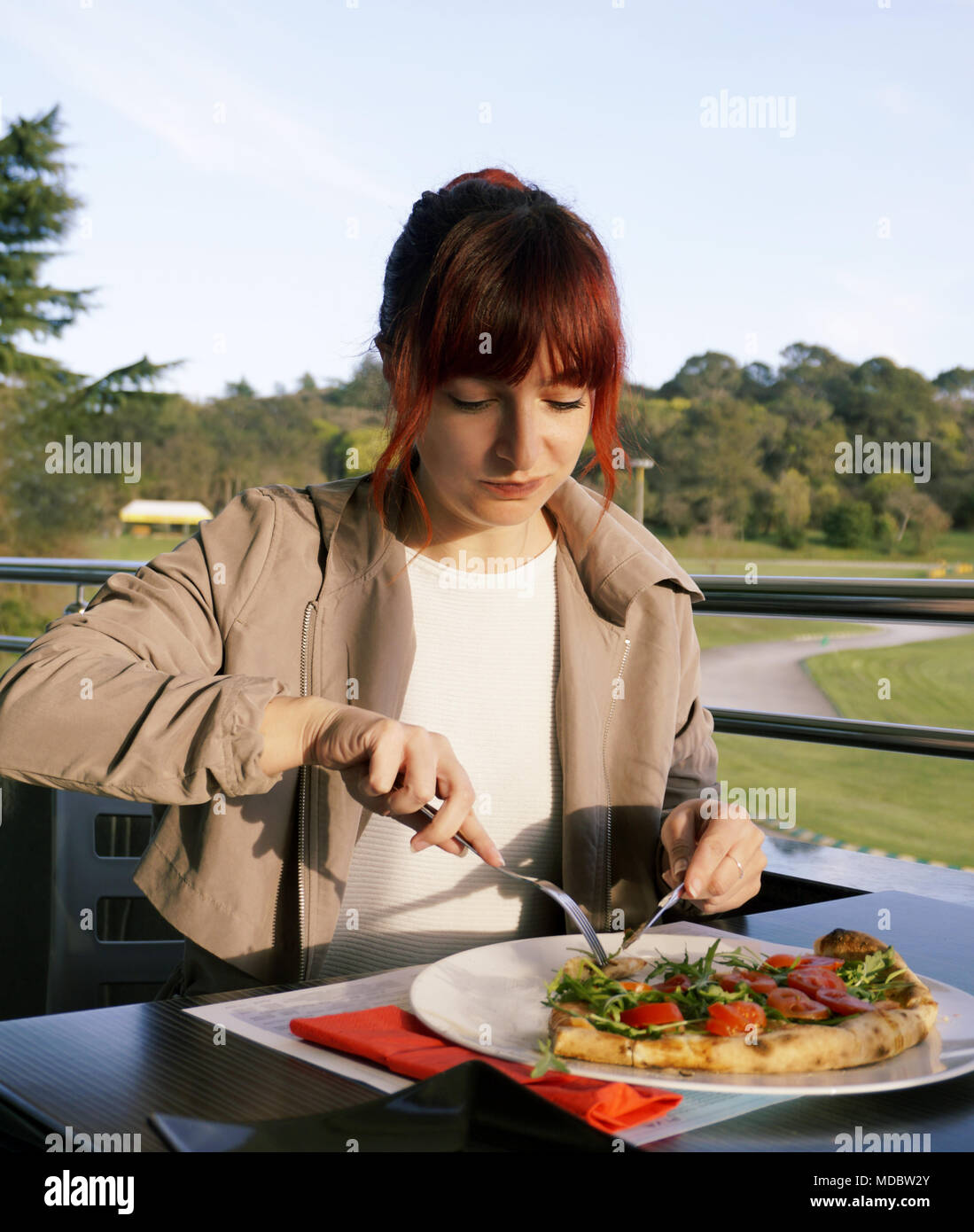 Redhead young woman eating pizza with fork and knife in the restaurant. Portrait of a young nice woman eating alone outside Stock Photo