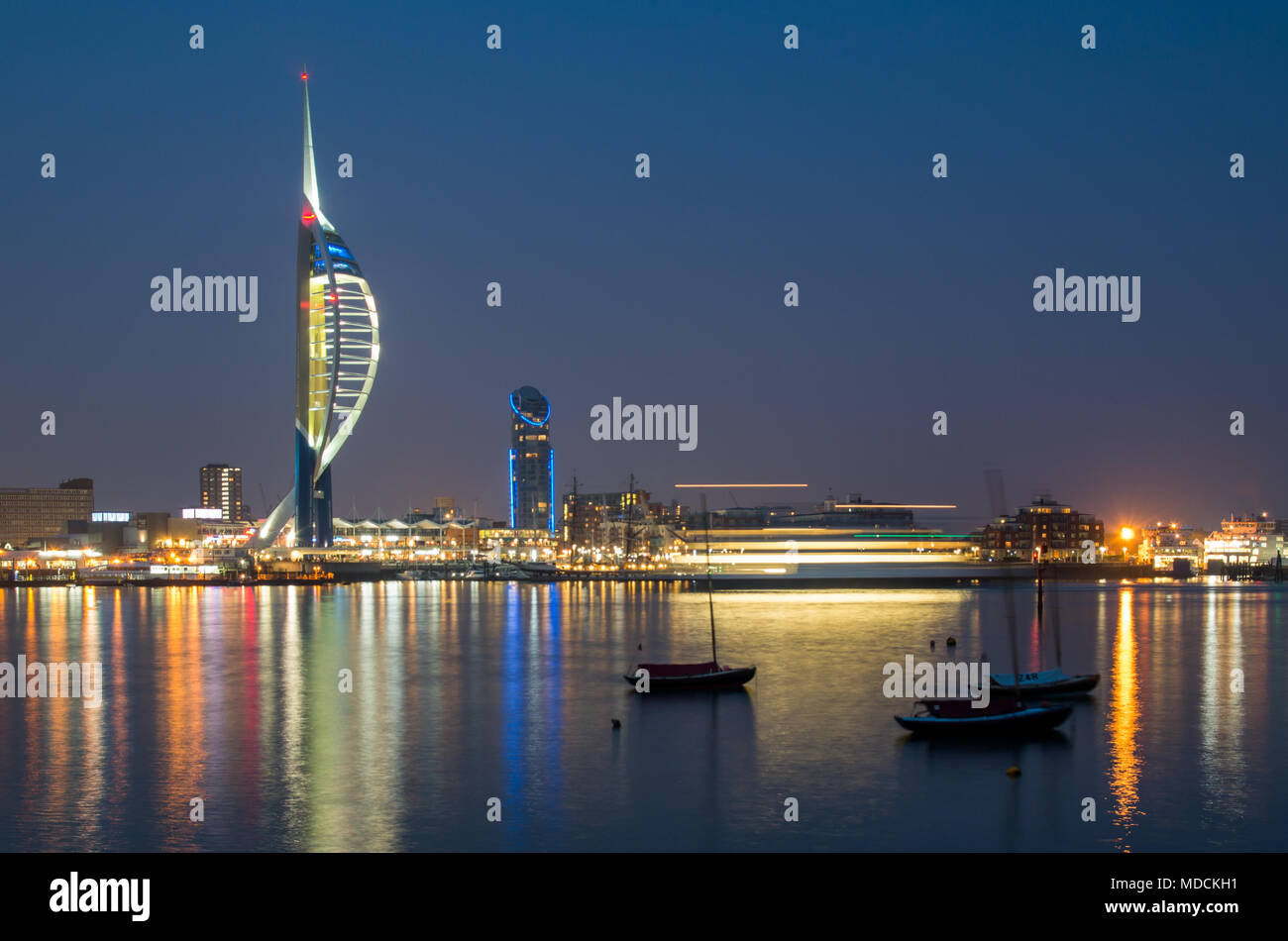 A long exposure night shot of Portsmouth harbour with few boats in the foreground, blurred shape of a passing ferry and illuminated Spinnaker tower. Stock Photo