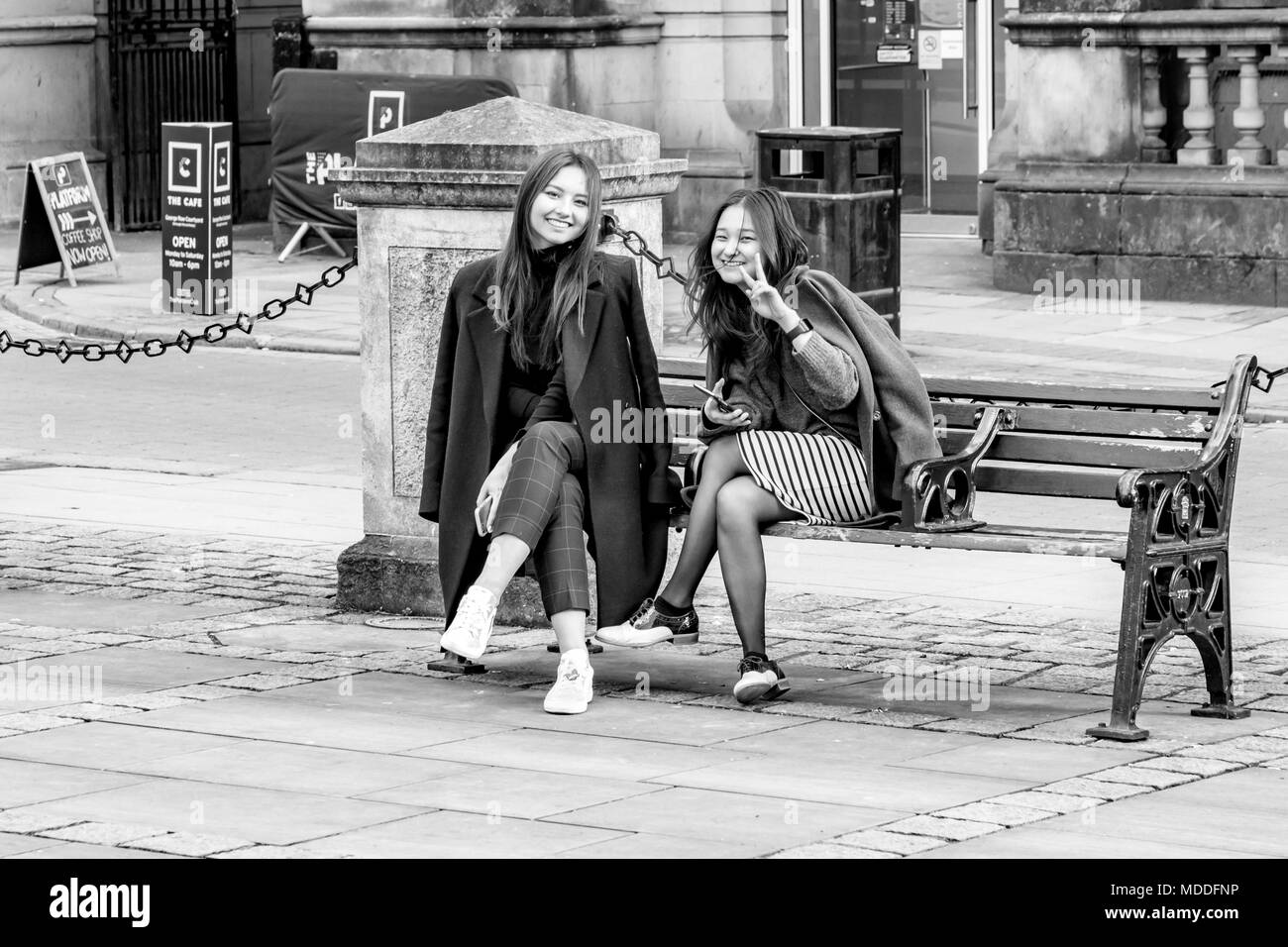 Two Oriental female friends from Northampton college sitting on a wrought Iron and wooden bench at the front of All Saints Church, Northampton town ce Stock Photo