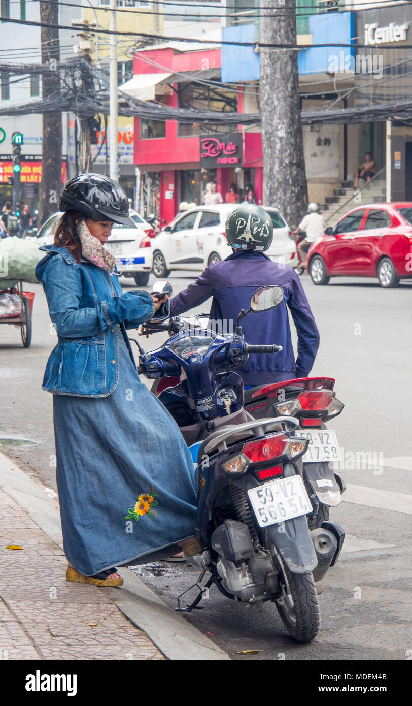 A Vietnamese woman wearing denim jacket, dress and gloves, using her mobile phone by her motorbike, in Ho Chi Minh City, Vietnam. Stock Photo