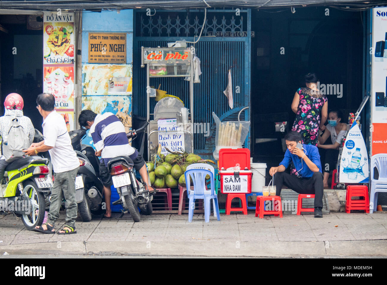 Motorcyclists parking their motorbikes on the pavement, and a Vietnamese man sitting on a plastic stool talking on his mobile phone and having a drink. Stock Photo