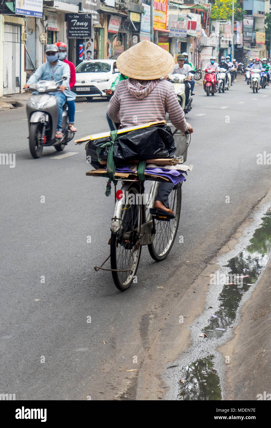 A Vietnamese female cyclist wearing a conical hat riding her bicycle on a street in Ho Chi Minh City, Vietnam. Stock Photo