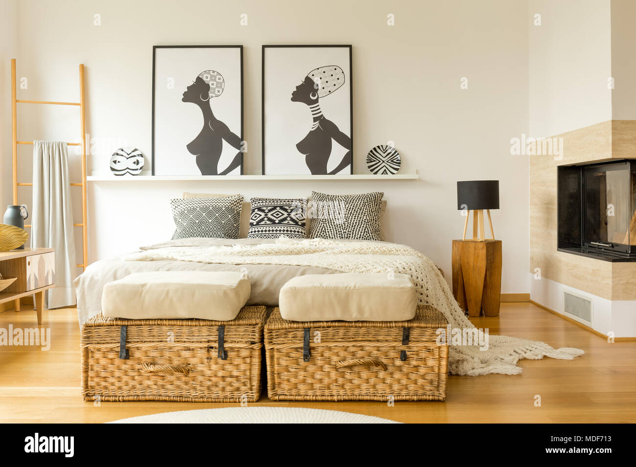 African posters, wicker boxes, fireplace and double bed with patterned pillows in a boho bedroom interior Stock Photo